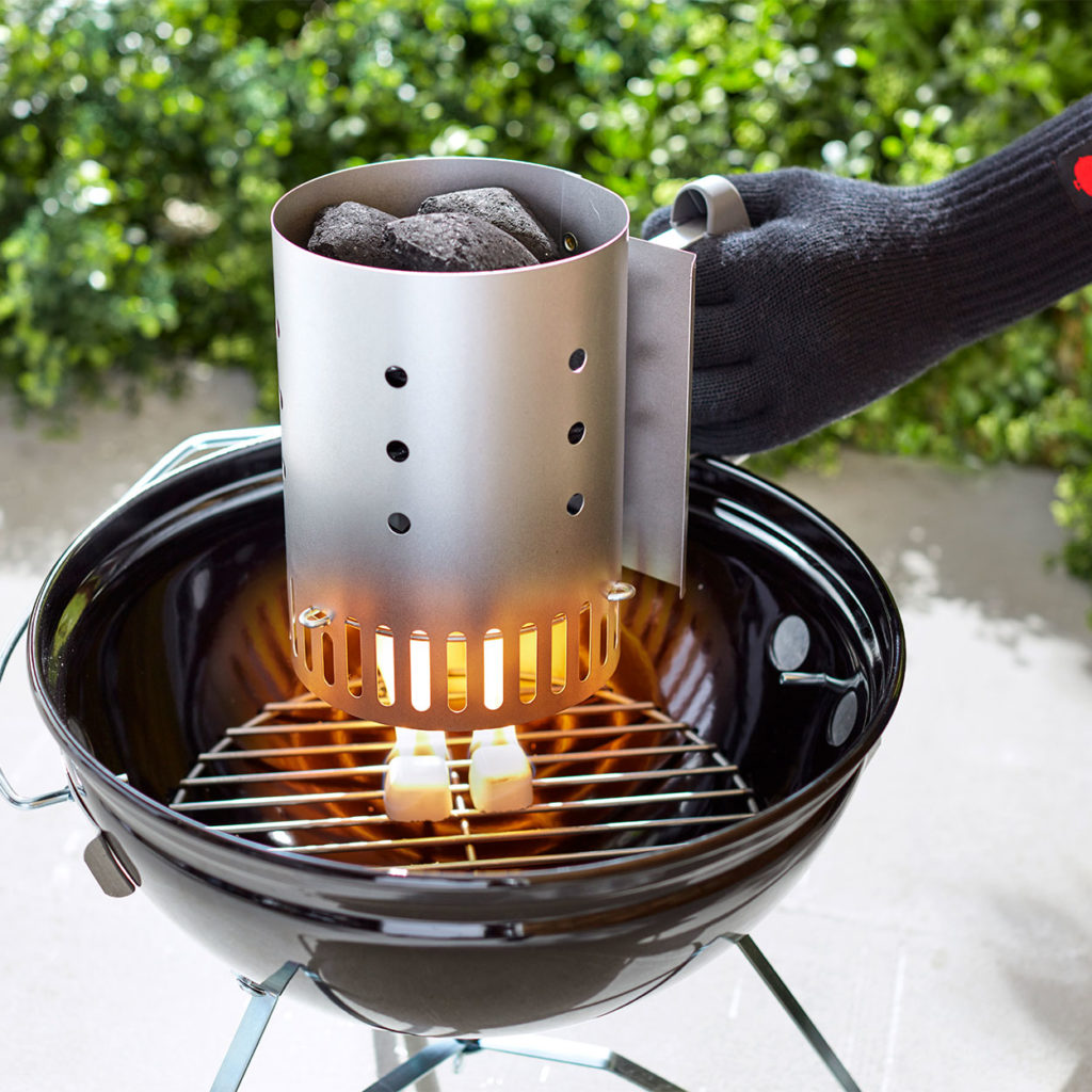 hand with glove hold a chimney starter filled with charcoal over lit cubes in bbq