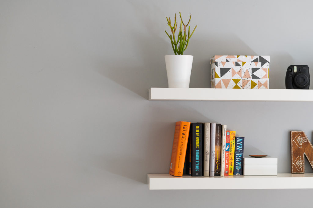 Two white wall shelves with books and plants placed on top of them 