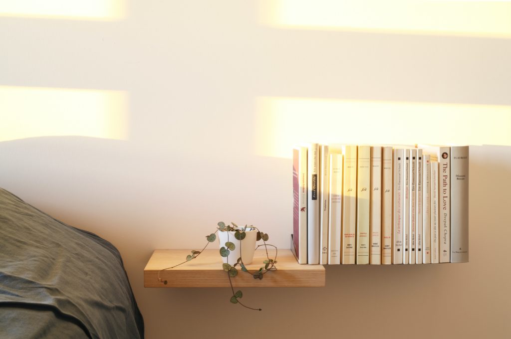 A floating wooden shelf beside a bed with books and a plant placed on it