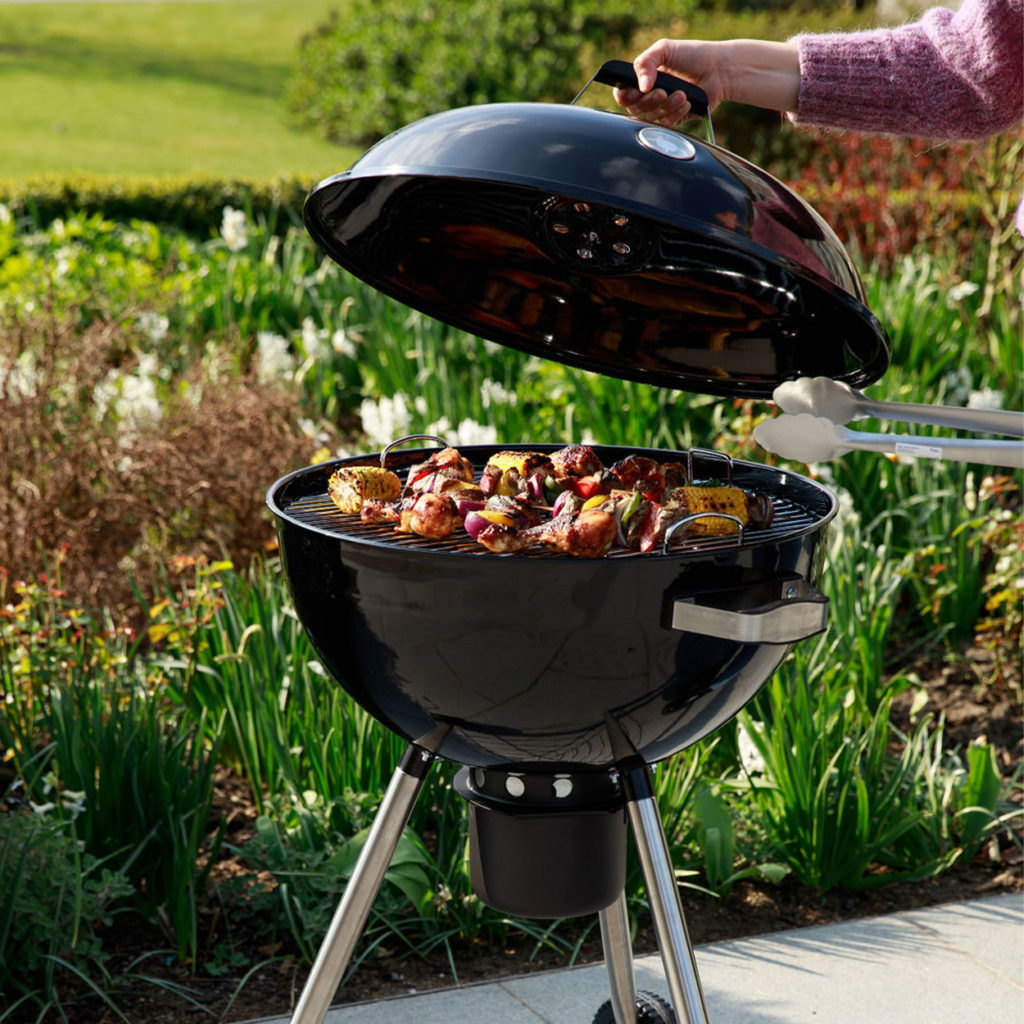 Hand holding tongs and opening charcoal BBQ with food cooking